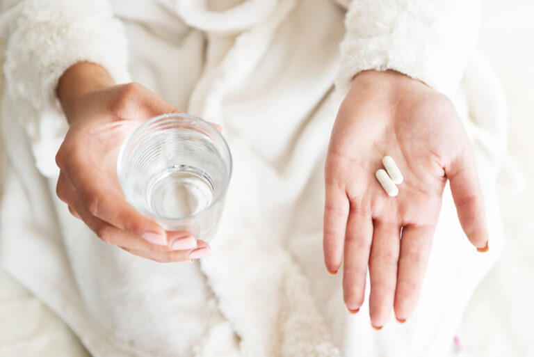 Close up of woman's hands holding pills in one hand and glass of water in other. She's wondering Lexapro vs Prozac.