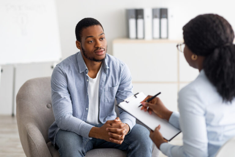 Man sitting in chair with hands folded in lap, talking to medical professional with clipboard