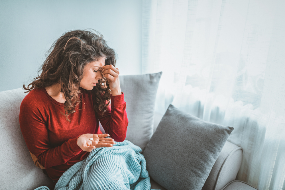 Upset young woman sitting on couch, eyes closed, hand pinching forehead with pill in other hand